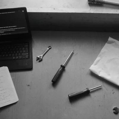 Mesa de trabajo con herramientas mecánicas reales junto a un portátil abierto en blanco y negro.