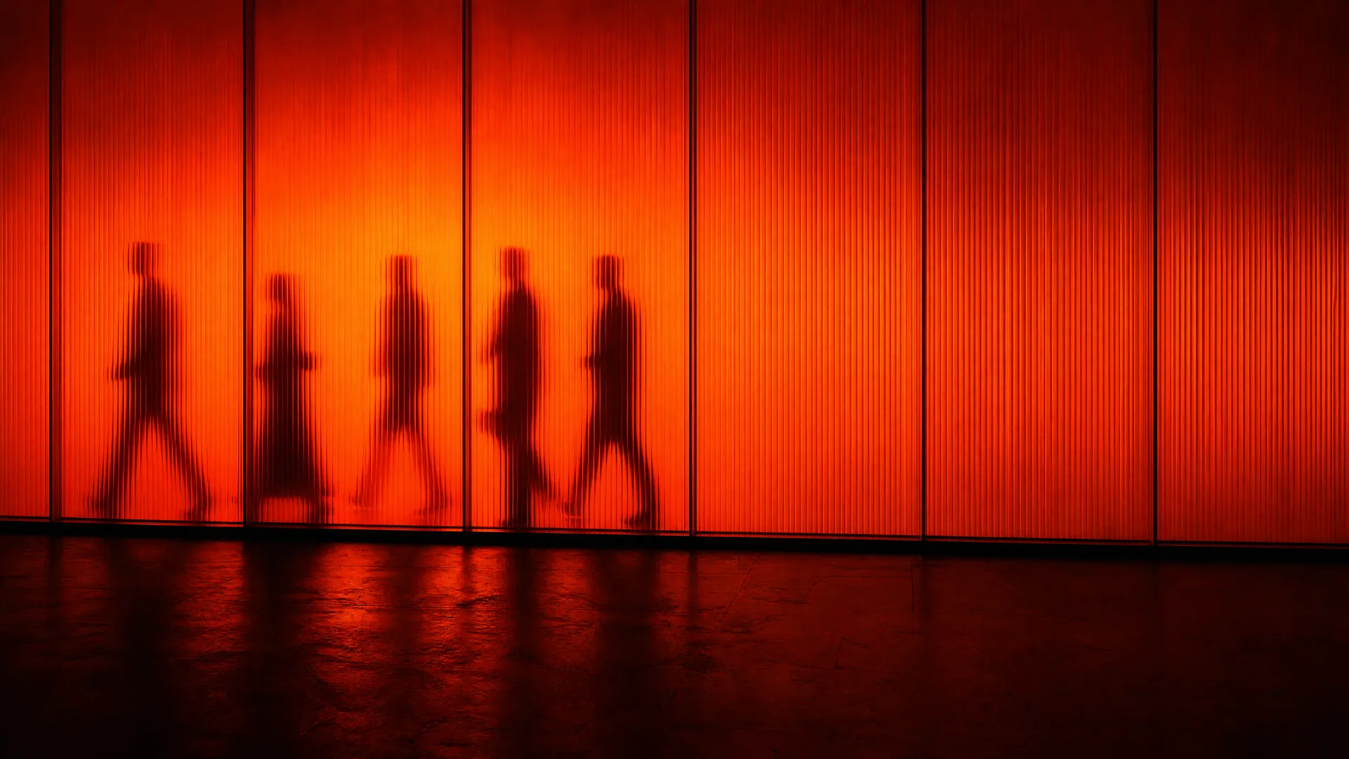 Un muro de vidrio acanalado retroiluminado en naranja con varias siluetas de personas pasando por detrás
