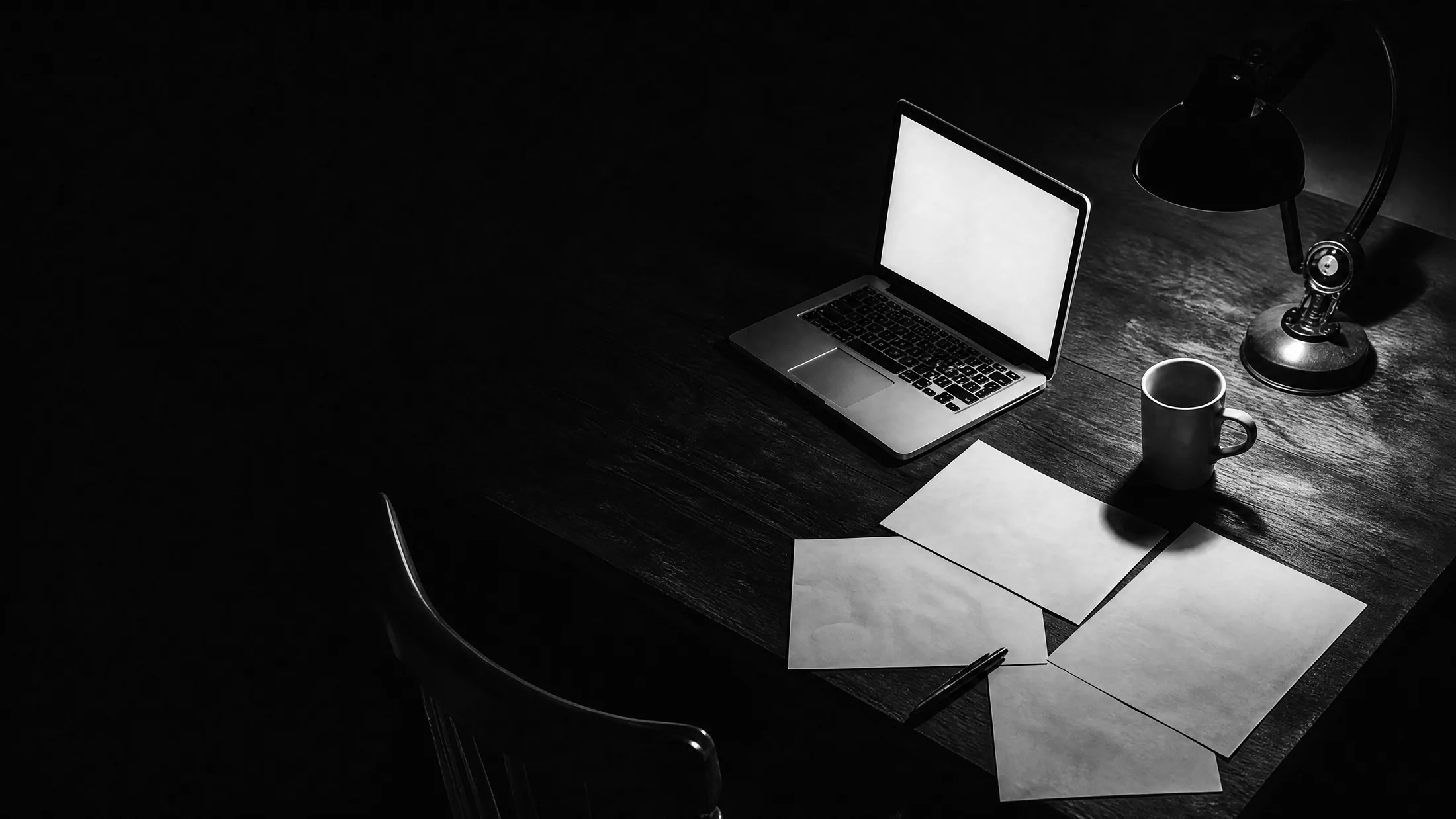 Una mesa de trabajo de noche con un portátil abierto, folios sin escribir y un bolígrafo, iluminada solo por la pantalla y un flexo